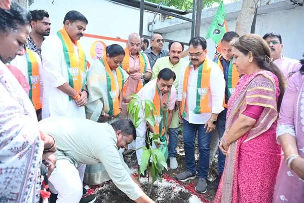 मैदान पर लोग गड्ढे में पौधा लगा रहे हैं, आसपास माला पहने जनप्रतिनिधि खड़े हैं